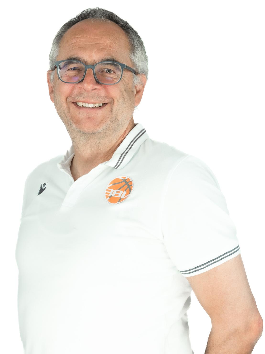 Portrait eines l&auml;chelnden Mannes mit Brille, der ein wei&szlig;es Poloshirt tr&auml;gt und vor einem hellen Hintergrund steht.