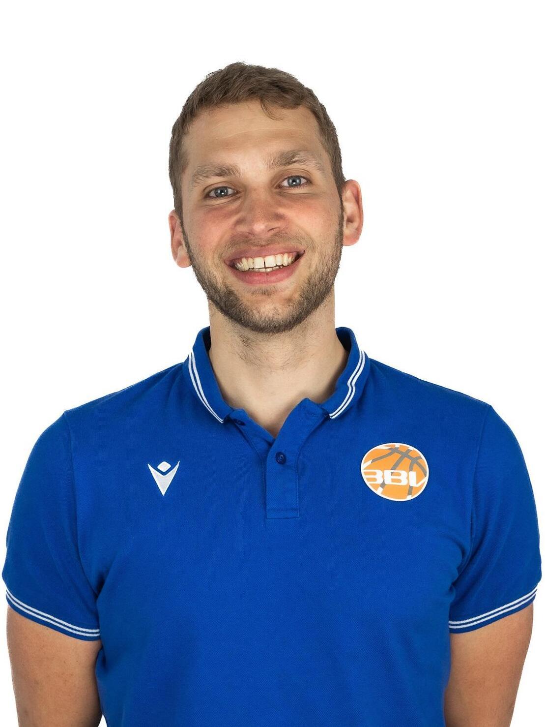 L&auml;chelnder Mann mit kurzer Frisur, tr&auml;gt ein blaues Poloshirt mit einem orangefarbenen Basketball-Logo.