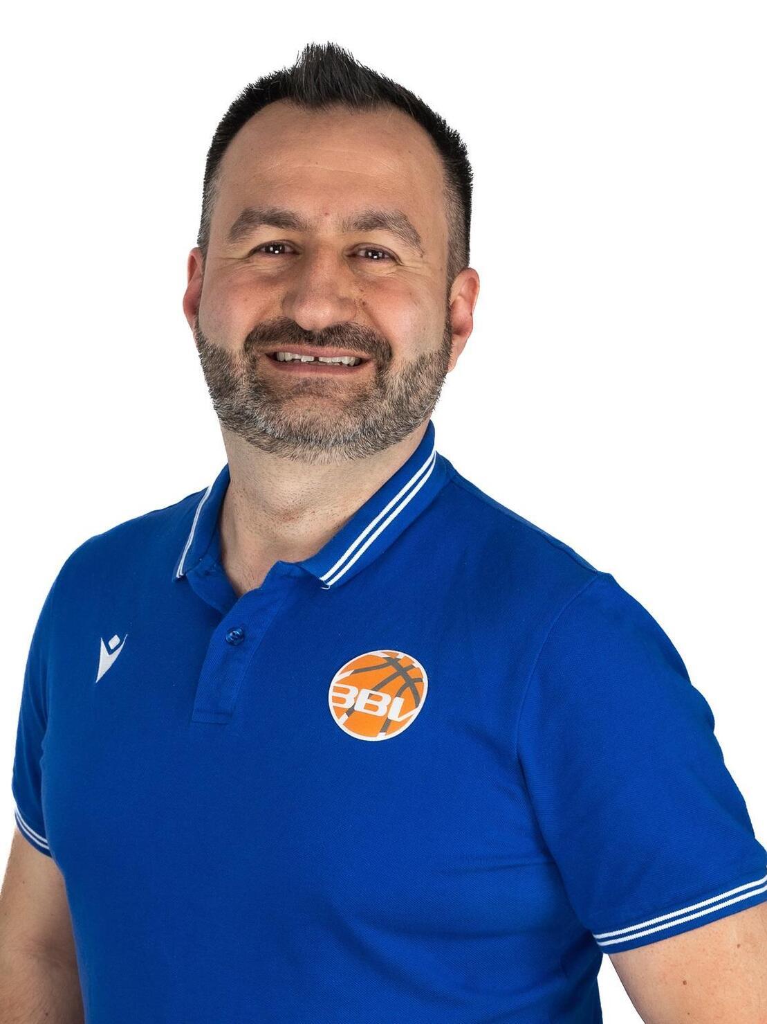 L&auml;chelnder Mann mit kurzer Frisur und Vollbart, tr&auml;gt ein blaues Polo mit Logo, vor neutralem Hintergrund.