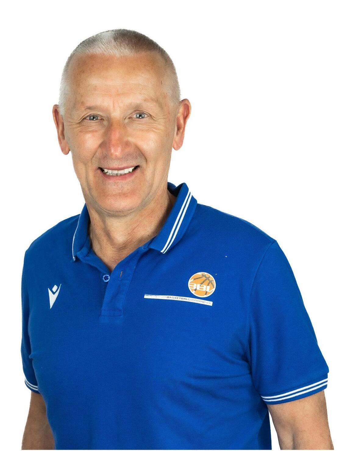 L&auml;chelnder Mann mit kurzen grauen Haaren in blauer Polo-Shirt vor wei&szlig;em Hintergrund.