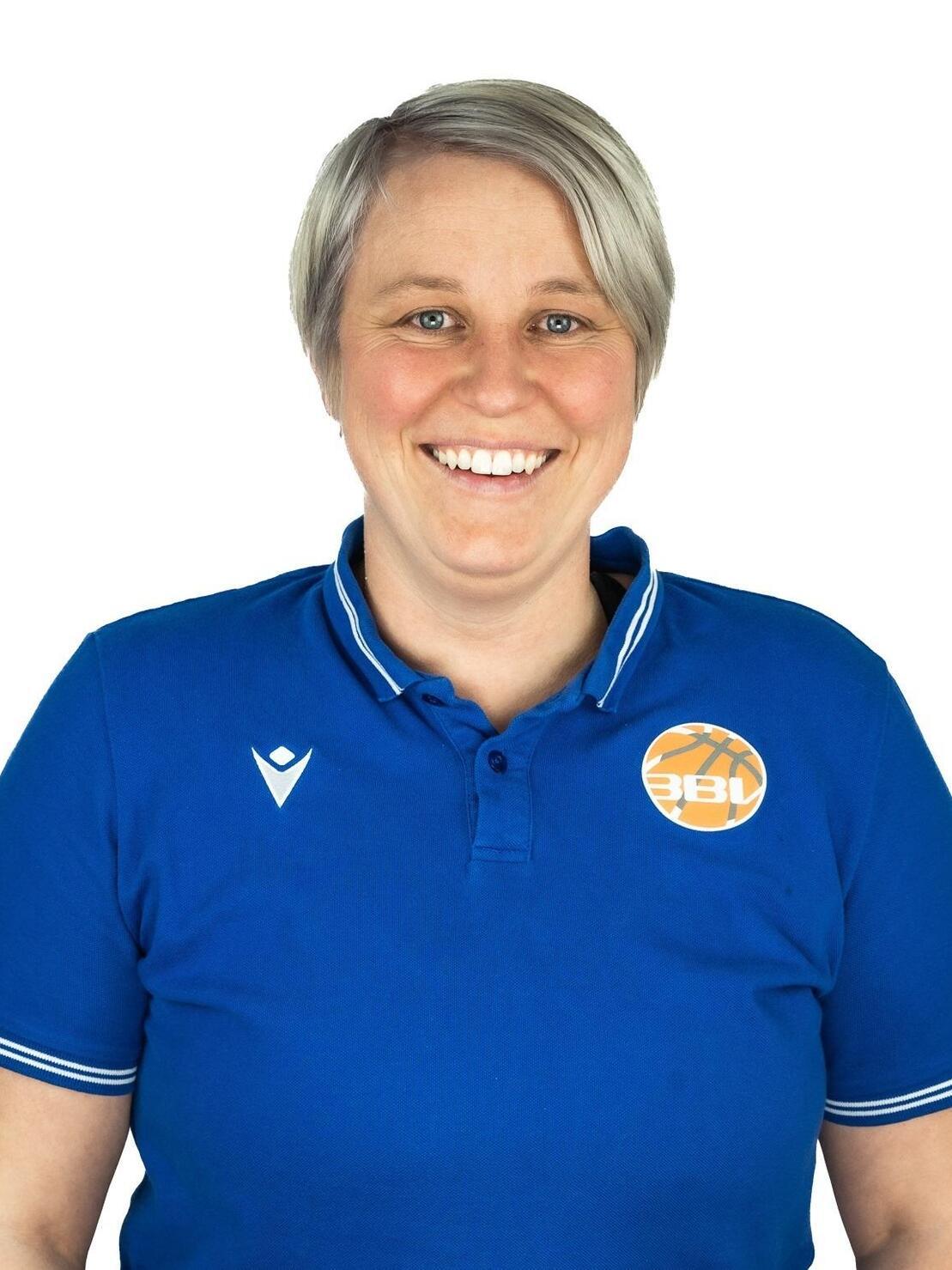 Eine l&auml;chelnde Frau mit kurzem, grauem Haar tr&auml;gt ein blaues Poloshirt mit einem Basketball-Logo.